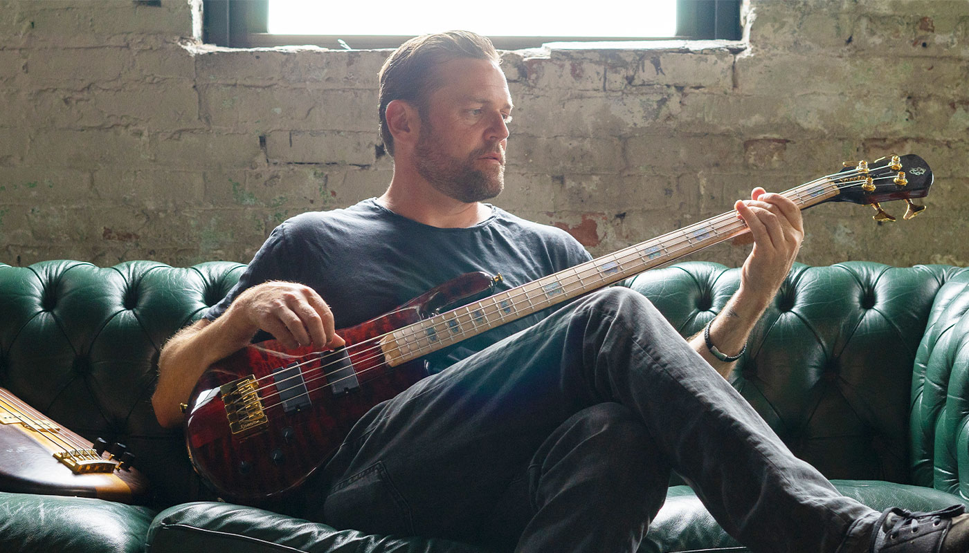 Man playing a Spector bass and sitting on a vintage leather couch in front of a worn concrete wall